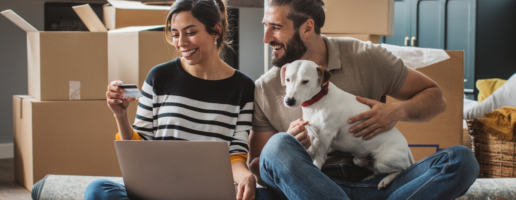 A smiling couple sitting on the floor of their new pet-friendly apartment surrounded by moving boxes, using a laptop and credit card with their dog.