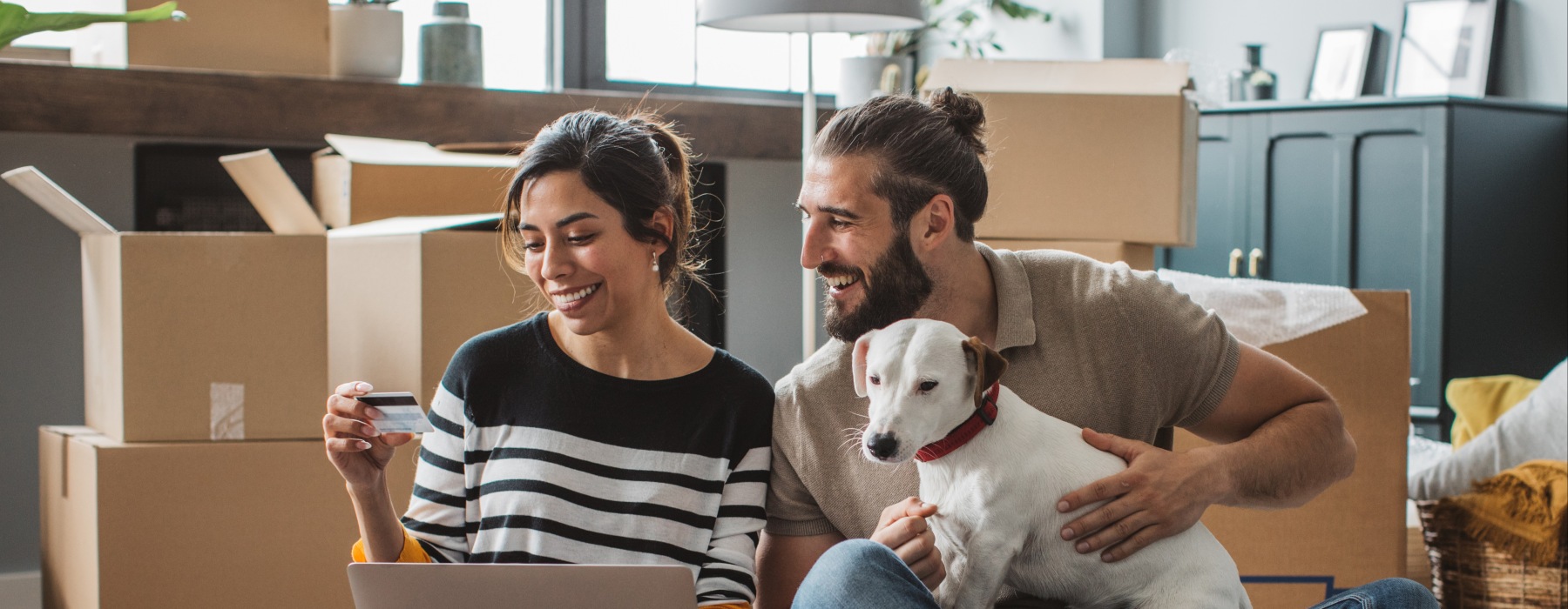 A smiling couple sitting on the floor of their new pet-friendly apartment surrounded by moving boxes, using a laptop and credit card with their dog.